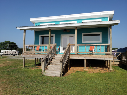 A bright turquoise house with a porch, steps, and orange chairs, set in a grassy area under a clear blue sky.