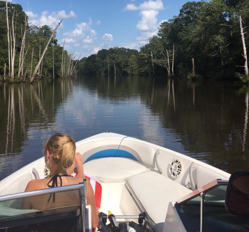 A person sits in a boat on a calm river, surrounded by trees and blue skies, enjoying a peaceful day on the water.