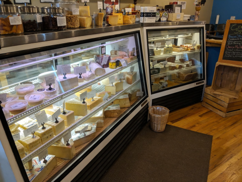 Display case filled with various types of cheese, showcasing a variety of shapes and labels in a cozy shop setting.