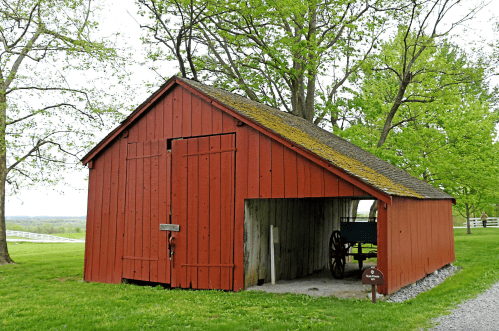 A red barn with a mossy roof, set in a grassy area with trees and a gravel path nearby.