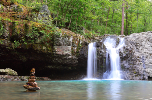 A serene waterfall cascades into a clear pool, with a stack of stones in the foreground surrounded by lush greenery.
