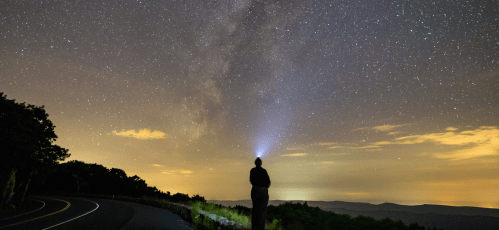 A person stands on a road at night, pointing a flashlight at a starry sky filled with the Milky Way.