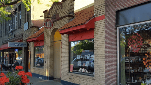 A brick storefront with a red awning, featuring large windows and a logo, surrounded by flowers and nearby shops.