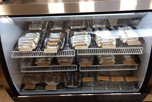 A display case filled with various cannoli flavors, including chocolate chip, peanut butter, and strawberry, labeled on trays.