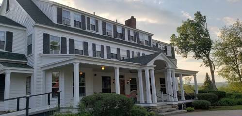 A large white house with multiple windows and a porch, surrounded by greenery under a cloudy sky.