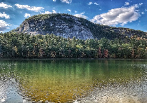 A serene lake reflects a rocky mountain surrounded by lush trees and colorful autumn foliage under a blue sky.