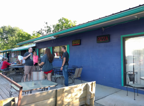People chatting outside a colorful building with signs for "Bar" and "Pizza," surrounded by outdoor seating.