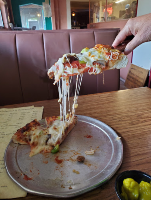 A hand lifts a cheesy slice of pizza, showcasing melted cheese and toppings, with a plate and condiments in the background.
