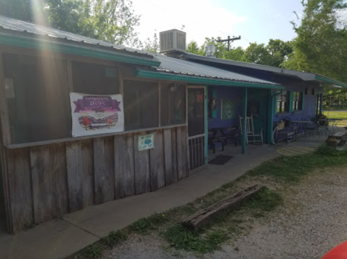 A rustic building with a wooden exterior, featuring a colorful sign and outdoor seating in a green, sunny area.