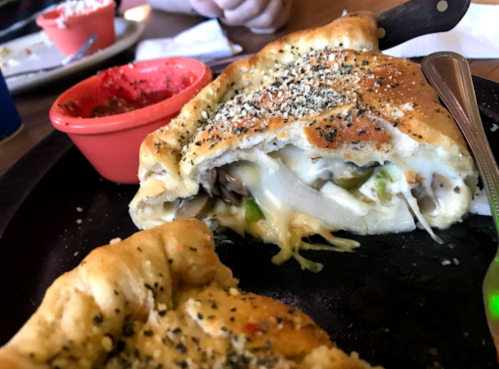 A close-up of a stuffed calzone with melted cheese and vegetables, served with marinara sauce on the side.
