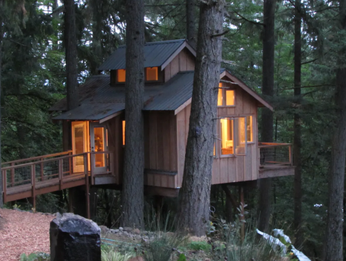 A cozy treehouse nestled among tall trees, featuring large windows and a wooden deck, illuminated at dusk.