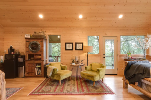 Cozy cabin interior with wooden walls, two green chairs, a coffee table, and large windows overlooking nature.