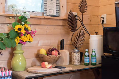 A cozy kitchen corner with flowers, fruit, a cutting board, and sparkling water bottles on a wooden table.