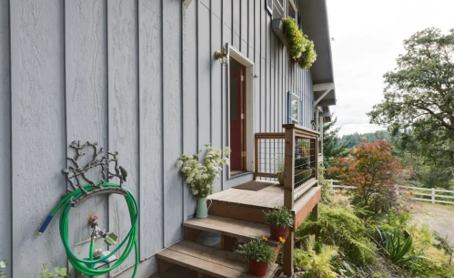 A wooden staircase leads to a gray house entrance, adorned with plants and a garden hose on the side.