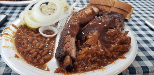 A plate of barbecue ribs, pulled pork, baked beans, pickles, onions, and slices of bread on a checkered tablecloth.