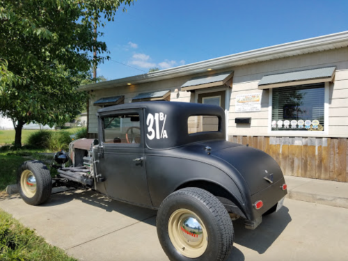 A vintage black car with a matte finish parked outside a building with a sign and several awards displayed.