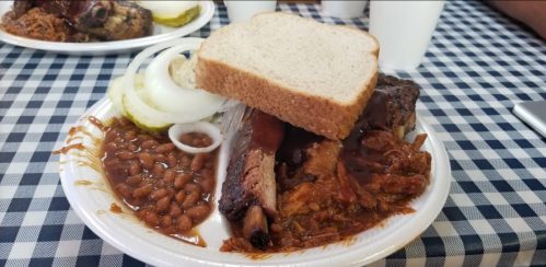 A plate of barbecue ribs with bread, onions, and a side of baked beans on a checkered tablecloth.