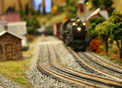 A model train approaches on a winding track surrounded by miniature trees and buildings in a scenic landscape.