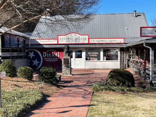 A rustic general store with a Tennessee flag, brick pathway, and a statue in front, set against a clear blue sky.