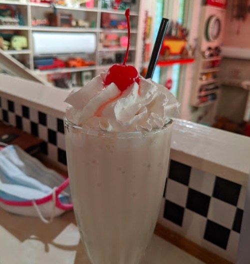 A tall glass of milkshake topped with whipped cream and a cherry, with two straws, set against a colorful background.