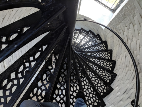 A spiral staircase with intricate black metalwork, viewed from above, showing the steps and a person's foot.