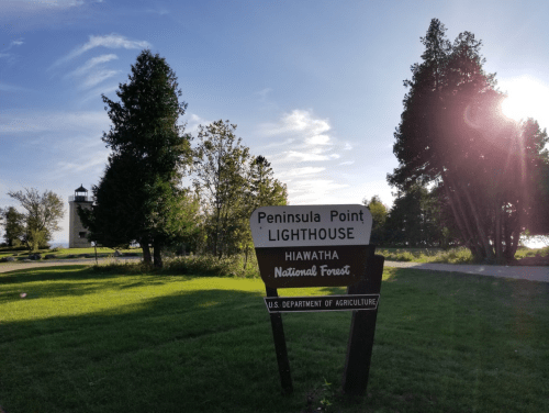 Sign for Peninsula Point Lighthouse in Hiawatha National Forest, with trees and sunlight in the background.