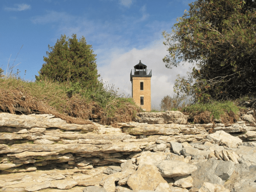 A lighthouse stands on a rocky shore, surrounded by greenery under a partly cloudy sky.
