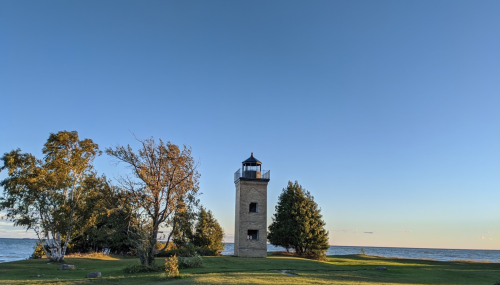 A lighthouse stands on a grassy shore, surrounded by trees, with a clear blue sky and calm water in the background.