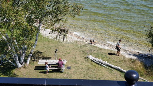 Aerial view of a lakeside scene with people near a picnic table, a child playing, and a man with a camera.