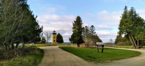 A scenic view of a lighthouse tower surrounded by trees, with a signpost and a winding dirt road leading to it.