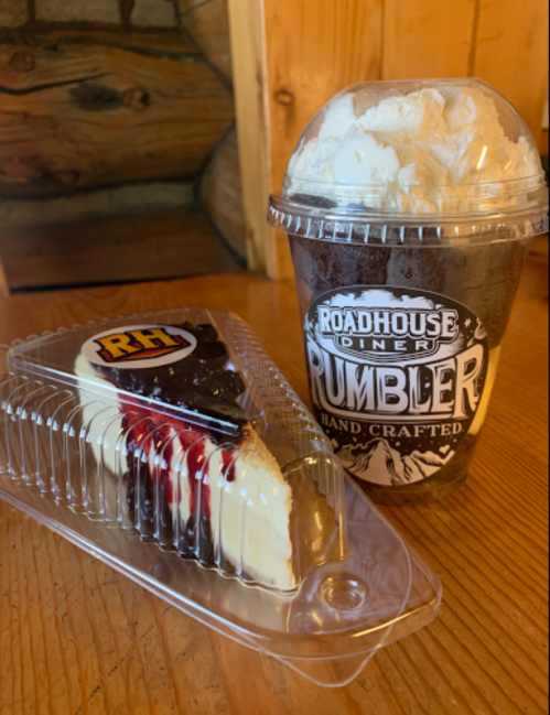 A slice of cheesecake in a container next to a frosty drink topped with whipped cream, on a wooden table.