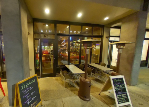 Exterior view of a restaurant with outdoor seating, chalkboard menus, and warm lighting in the evening.