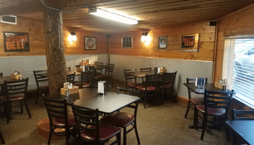 Cozy restaurant interior with wooden walls, tables, and chairs, featuring a central tree and warm lighting.