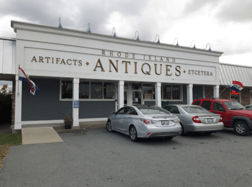 Exterior of a store labeled "Rhode Island Antiques" with a gray car parked in front and a red vehicle nearby.