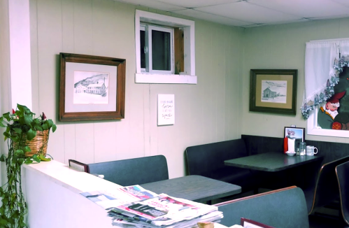 Cozy diner interior with framed artwork, a window, and a small table set for dining. Newspapers are stacked on the counter.