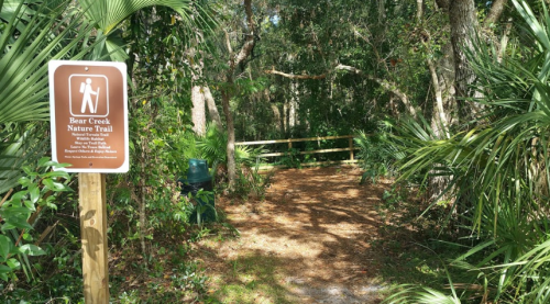 A wooded path leading into Bear Creek Nature Trail, marked by a sign and surrounded by lush greenery.