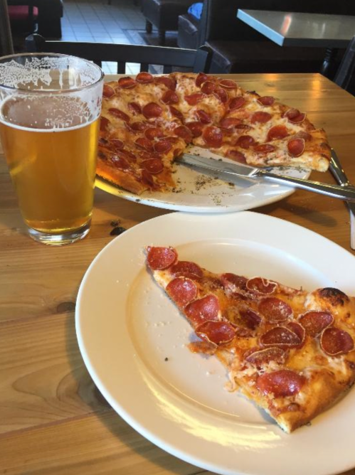 A slice of pepperoni pizza on a plate next to a full pizza and a glass of beer on a wooden table.