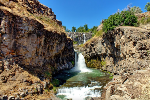 A scenic waterfall cascading into a rocky gorge, surrounded by greenery and clear blue skies.