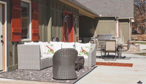 A cozy outdoor patio with a white sofa, wicker chair, and a grill, surrounded by green walls and sunny weather.