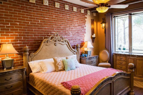 Cozy bedroom with a brick wall, wooden furniture, a large bed, and warm lighting from lamps and a ceiling fan.