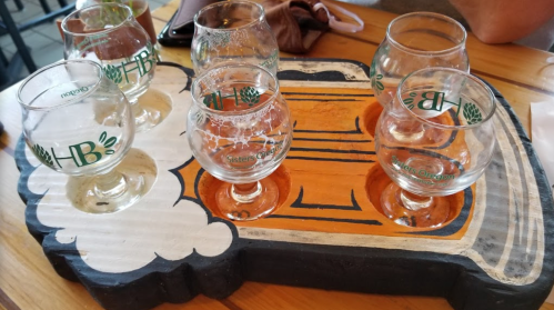 A wooden tray with six small glasses, some filled with beer, arranged on a table.