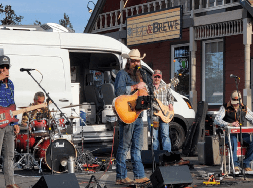 A country band performs outdoors, with a singer holding a guitar and musicians on drums, bass, and keyboard.