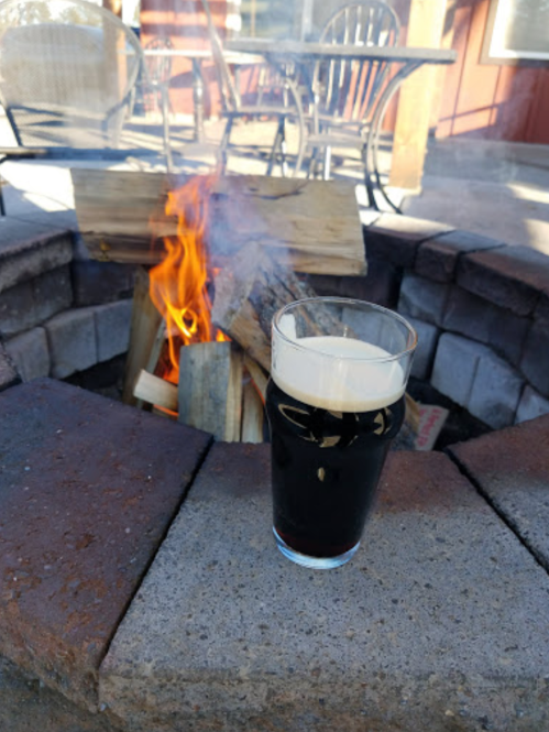 A glass of dark beer sits on a stone fire pit, with flames and logs burning in the background.
