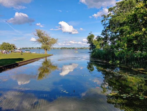 A serene lake scene with blue skies, fluffy clouds, and reflections on the water, surrounded by trees and greenery.