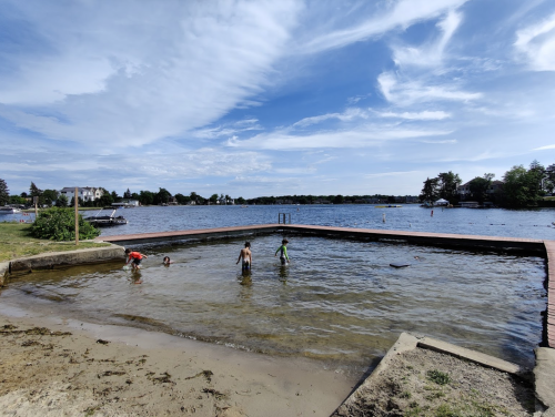 Children playing in shallow water at a lakeside beach, with boats and trees in the background under a blue sky.