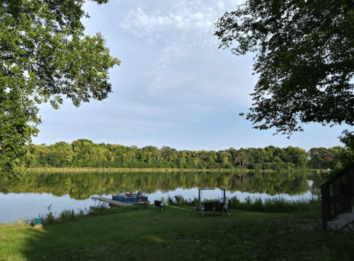A serene lake surrounded by trees, reflecting the sky, with a small dock and seating area on the grassy shore.