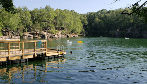 A serene lake surrounded by trees, with people swimming and floating on inflatable toys near a wooden dock.