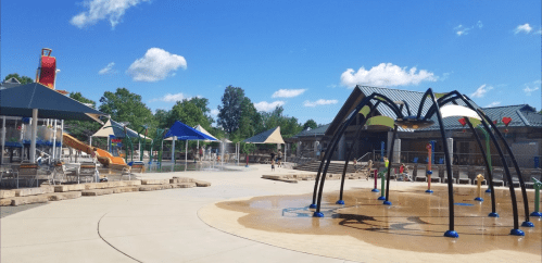 A sunny water park scene featuring slides, splash pads, and shaded seating areas. Families enjoy the outdoor space.