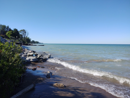 A serene lakeshore scene with gentle waves, rocky shoreline, and clear blue skies.