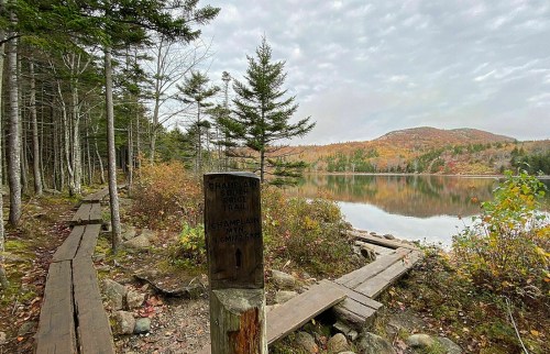 A wooden sign marks the start of a hiking trail by a calm lake, surrounded by autumn foliage and mountains in the background.